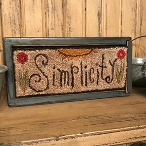 May include: A rectangular wooden sign with the word "Simplicity" embroidered in dark brown thread. The sign features a beige background with a sun and flowers. The frame is painted blue.