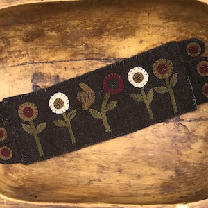 Peut inclure: Textile rectangulaire marron foncé avec des fleurs brodées, présenté dans un bol en bois. Les fleurs ont des centres blancs, rouges et bruns avec des tiges et des feuilles vertes. Le bol est en bois brun clair, avec des traces d'usure.