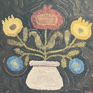 May include: A rug hooking depicting a bouquet of flowers in a white pot. The flowers are yellow, red, and blue. The background is a dark brown.