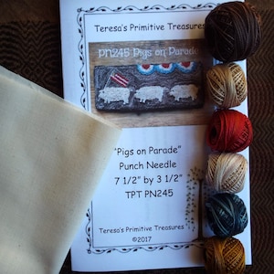 May include: A craft kit labelled "Pigs on Parade" by Teresa's Primitive Treasures. The kit contains a printed pattern, a piece of fabric, and several balls of yarn in various colours. The pattern measures 19 cm by 9 cm.