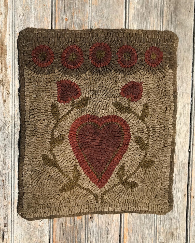 May include: A brown and beige hooked rug with a red heart in the center surrounded by green leaves. The rug has a border of red and brown flowers.