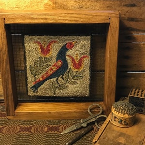 Puede incluir: Una obra de arte textil enmarcada que presenta un pájaro azul con detalles rojos, posado entre flores rojas y hojas verdes. La obra de arte está en un marco de madera con una malla de alambre. También se ven herramientas de costura vintage.