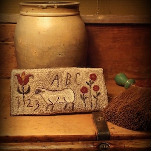 Op de afbeelding: Een rechthoekig, beige tapijt met een schaap, bloemen en de letters "ABC" en de cijfers "123". Achter het tapijt staat een grote, beige stenen pot. Rechts bevinden zich een klein groen voorwerp en een bezem.