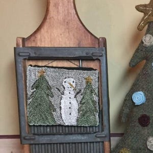 May include: A rustic wooden and metal Christmas decoration featuring a framed rug with a snowman and two green Christmas trees. The frame is set on a wooden base with a handle. A felt Christmas tree is visible in the background.