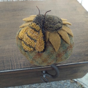 May include: A felt bee with yellow and black stripes sits on a felt sunflower with brown and green petals. The bee and sunflower are handmade and have a rustic, vintage look.
