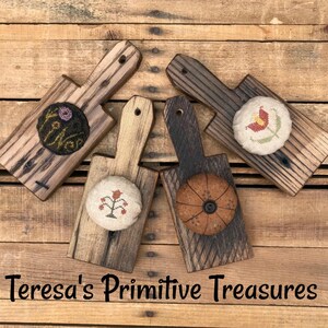 May include: Four wooden cutting boards with attached decorative pincushions. The pincushions feature embroidered floral designs in various colors. The boards are made of weathered wood and are arranged on a wooden surface. The text "Teresa's Primitive Treasures" is at the bottom.
