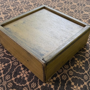 May include: A square wooden box with a lid. The box is painted in a light olive green color with darker green accents around the edges. The wood grain is visible on the surface.