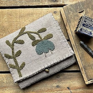 Könnte beinhalten: Eine kleine graue Stofftasche mit einem Klappverschluss. Die Tasche ist mit einer grünen Filzapplikation einer stilisierten Blume mit einem blauen Zentrum verziert.