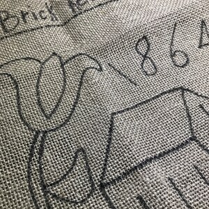 May include: Close-up of a gray burlap fabric with black sketched outlines of a floral design and a house. The text "Brick Red Saltbox" and the numbers "1864" are also visible in black ink.
