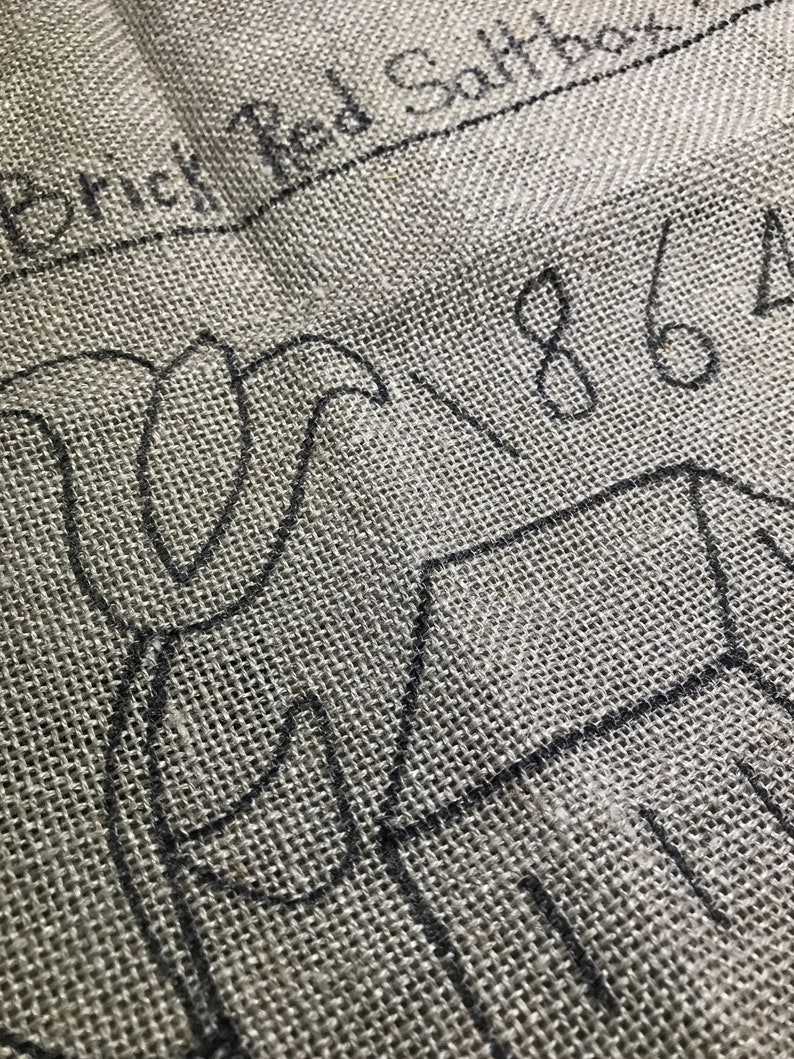 May include: Close-up of a gray burlap fabric with black sketched outlines of a floral design and a house. The text "Brick Red Saltbox" and the numbers "1864" are also visible in black ink.