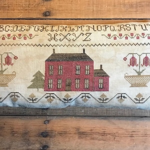 May include: A white pillow with a red house, green trees, and red flowers embroidered on it. The pillow has the alphabet embroidered at the top and a border of brown and gold.