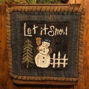 May include: A framed, square, winter-themed wall hanging with the words "Let it Snow" in white script. The image features a snowman, a Christmas tree, and a fence. The frame is brown with a stitched border.