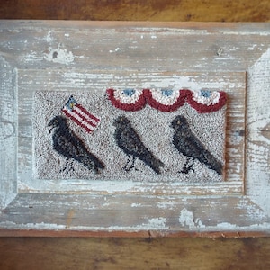 Puede incluir: Una pieza de punto de aguja enmarcada con tres pájaros negros y una bandera estadounidense roja, blanca y azul. Los pájaros están alineados en una fila y la bandera está en el fondo. Hay banderines rojos, blancos y azules sobre los pájaros.