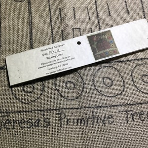 May include: A close-up of a burlap fabric with a drawn design of a house and circles. A tag reads "~Brick Red Saltbox~ Size 15x13 Backing Linen" with additional text and a small image. The fabric is tan.