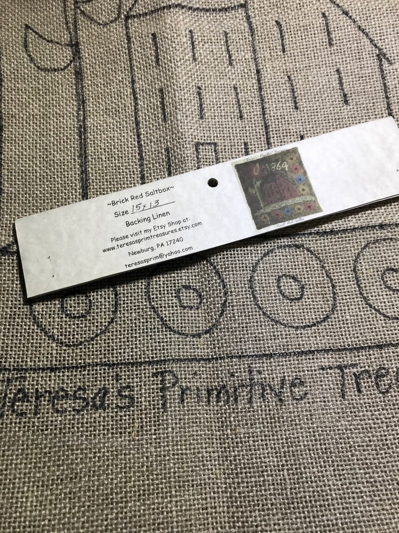 May include: A close-up of a burlap fabric with a drawn design of a house and circles. A tag reads "~Brick Red Saltbox~ Size 15x13 Backing Linen" with additional text and a small image. The fabric is tan.