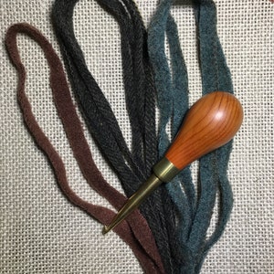 May include: A wooden handle rug hooking tool with a brass ferrule and hook, surrounded by strands of brown, dark gray, and teal wool strips. The items are arranged on a textured, light-colored surface.