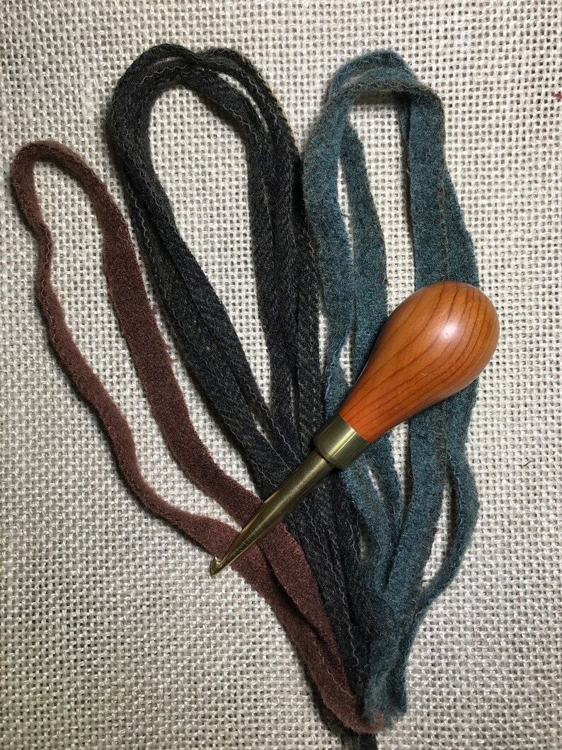 May include: A wooden handle rug hooking tool with a brass ferrule and hook, surrounded by strands of brown, dark gray, and teal wool strips. The items are arranged on a textured, light-colored surface.