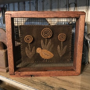 以下が含まれることがあります： 茶色の背景に茶色の鳥と黄色の芯を持つ茶色の花が3つ描かれた、額装された針仕事の作品。フレームは木でできており、ワイヤーメッシュの前面があります。