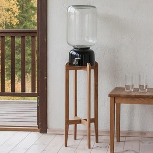 Peut inclure: Un distributeur de boissons en verre transparent avec une base noire et un robinet argenté, soutenu par un haut support en bois à quatre pieds. Le distributeur est placé à côté d'une petite table d'appoint en bois avec deux verres.
