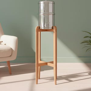 Op de afbeelding: Een roestvrijstalen waterfiltersysteem met een kraan, op een lichtbruine houten standaard. Het filter bestaat uit twee gestapelde cilindrische containers. De standaard heeft een eenvoudig ontwerp. De achtergrond is een lichtgroene muur en een plant in een pot.