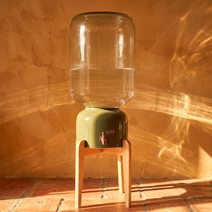 May include: A clear glass water dispenser with a large cylindrical reservoir sits atop a sage green ceramic base. The base features a metallic spigot and is supported by a light-colored wooden stand with four tapered legs, holding the unit upright.