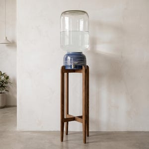 Puede incluir: Un dispensador de agua de vidrio transparente se asienta sobre una base de cerámica azul con patrones de líneas horizontales blancas. La base cuenta con un grifo de metal plateado y descansa sobre un soporte de madera alto de cuatro patas.