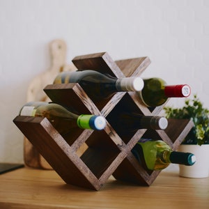 May include: A dark brown wooden wine rack with a criss-cross design holds six bottles of wine. The rack is on a light brown wooden surface.