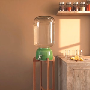 Op de afbeelding: Een grote waterdispenser van helder glas met een groene keramische voet en een zilverkleurige metalen kraan, ondersteund door een hoge houten standaard. De dispenser is gevuld met water en staat in een kamer met een houten aanrecht.