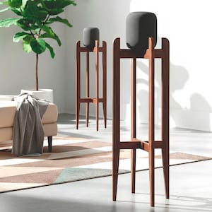 May include: Two dark grey, cylindrical speakers sit on tall, wooden stands. The stands are a rich brown colour and have a minimalist design with four legs and a cross-shaped support. The speakers appear to be made of a textured fabric.