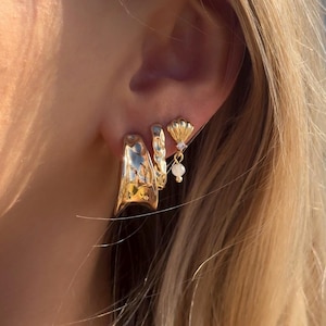 May include: Gold hoop earrings with a textured finish, layered with a smaller gold hoop earring and a gold charm with a white pearl.