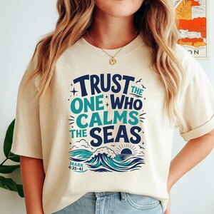 Może przedstawiać: Kremowy t-shirt z ciemnoniebieskim i morskim napisem "TRUST THE ONE WHO CALMS THE SEAS" nad grafiką przedstawiającą fale oceaniczne i wschodzące słońce. Napis "MARK 4:35-41" jest wydrukowany małymi literami pod ilustracją fal.