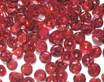 May include: A pile of red iridescent sequins, scattered on a white background. The sequins are round and have a slightly raised center.