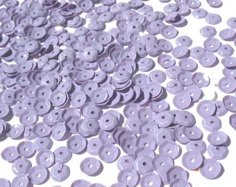 Könnte beinhalten: Eine Streuung von helllavendelfarbenen Pailletten, jede mit einem zentralen Loch und einem facettierten, geometrischen Design. Die Pailletten sind auf einer weißen Oberfläche verstreut und erzeugen einen strukturierten, dekorativen Effekt.