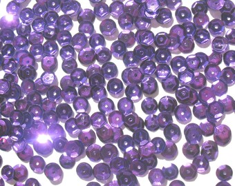 May include: A scattering of iridescent purple sequins on a white background. The sequins are round and have a slightly raised center.
