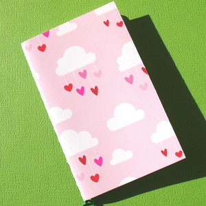 Peut inclure: Un carnet rose avec un motif de nuages et de cœurs. Les nuages sont blancs et les cœurs sont rouges, roses et blancs.