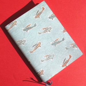 May include: A blue notebook cover with a repeating pattern of vintage aeroplanes in various colours. The aeroplanes are all facing the same direction and are arranged in a staggered pattern.