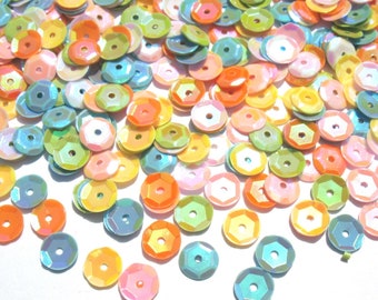Könnte beinhalten: Sortiment aus irisierenden Pailletten in verschiedenen Pastellfarben, darunter Rosa, Blau, Grün und Gelb. Die Pailletten sind auf einem weißen Hintergrund verstreut.