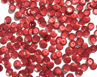 May include: A large quantity of red sequins, scattered on a white surface. The sequins are round and have a metallic sheen.