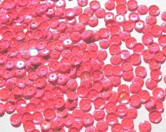 Könnte beinhalten: Eine Streuung von leuchtend pinkfarbenen Pailletten. Jede Paillette ist rund mit einem zentralen Loch und einem facettierten, sechseckigen Außenrand. Die Pailletten sind auf einer weißen Oberfläche verstreut und fangen das Licht ein.