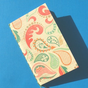 May include: A small, handmade notebook with a colourful paisley pattern in shades of green, orange, and pink. The notebook is closed and has a green string tied around it.