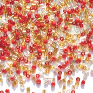 May include: A mix of red, gold, and clear glass seed beads. The beads are small and round, and they are scattered on a white surface.