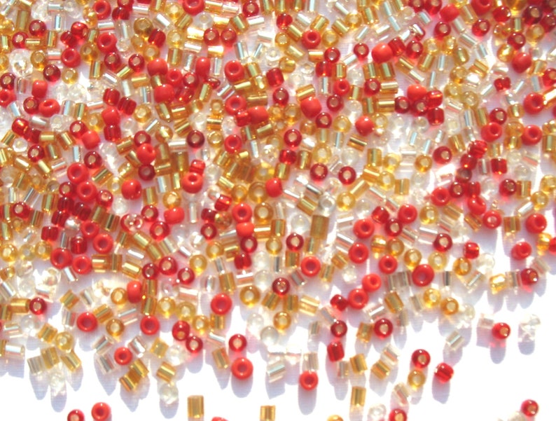 May include: A mix of red, gold, and clear glass seed beads. The beads are small and round, and they are scattered on a white surface.