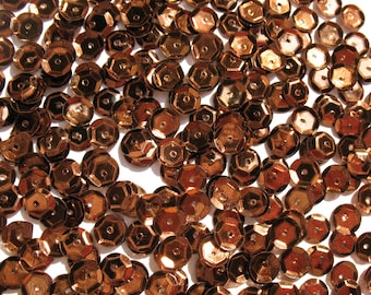 May include: A large quantity of copper-colored sequins, scattered on a white surface. The sequins are hexagonal in shape and have a metallic sheen.