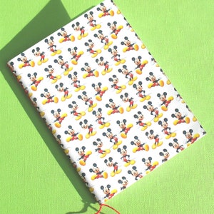 Puede incluir: Una libreta blanca con cierre de cordón rojo. La portada presenta un patrón repetido de Mickey Mouse en varias poses.