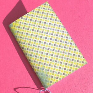 May include: A small, yellow, green and blue patterned notebook with a blue string closure. The notebook is lying on a pink surface.