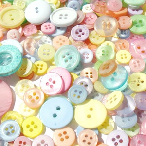 Op de afbeelding: Een verzameling pastelkleurige knopen in verschillende maten en vormen. De knopen zijn in tinten roze, geel, blauw, groen en wit. Sommige knopen hebben twee gaten, andere vier. De knopen zijn verspreid over een wit oppervlak.