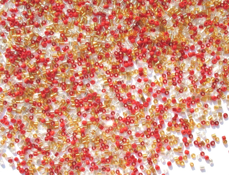 May include: A mix of red, gold, and clear glass seed beads. The beads are small and round, and they are scattered on a white background.