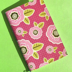 May include: A pink notebook with a floral pattern featuring pink flowers with brown centers and green leaves on a pink background with a white polka dot pattern.