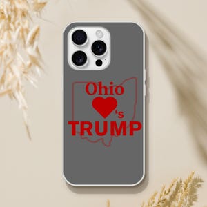 May include: A gray phone case with a white border. The case features a red outline of the state of Ohio, a red heart, and the words "Ohio's TRUMP" in red. The phone case is designed for a smartphone.