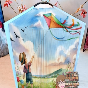 May include: A book sculpture of a child flying a kite in a blue sky with white clouds. The sculpture is made of pages from a book, and the kite is made of colorful paper. The sculpture is on a table with a stack of books in the background.